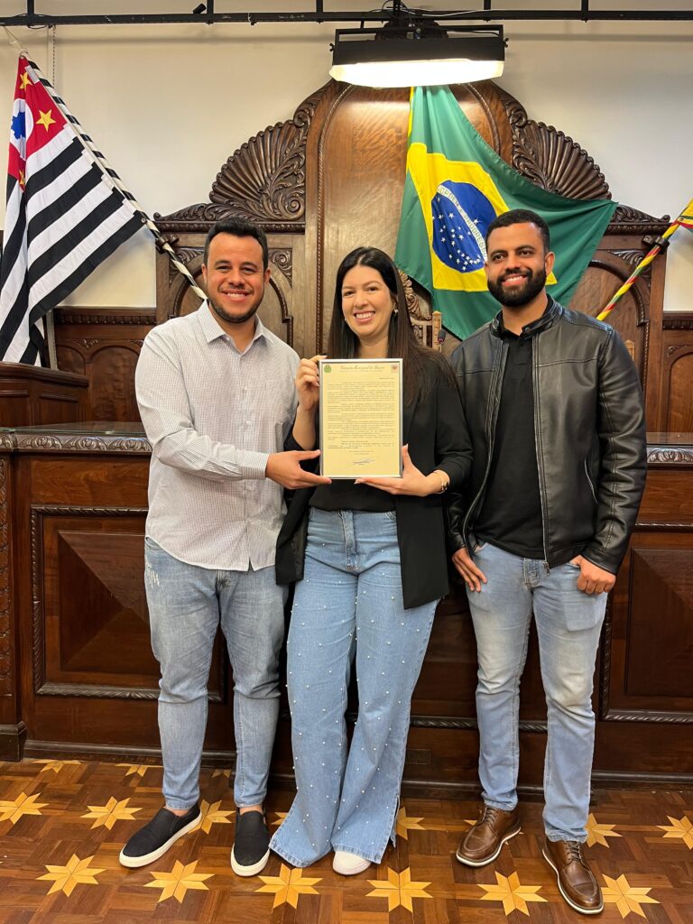 Três pessoas estão sorridentes em um ambiente interno, segurando uma placa ou certificado. Atrás delas estão um pódio de madeira, a bandeira do Brasil e a bandeira do estado de São Paulo - um evento oficial possivelmente celebrando a Instalação de ar condicionado em Agudos.